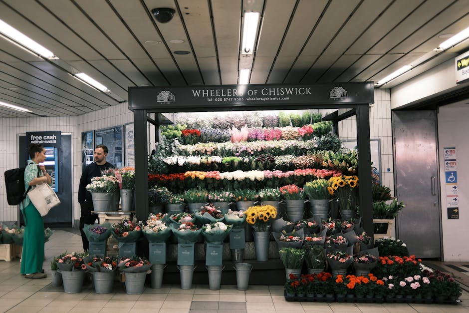 A floral display at Wheeler of Chiswick inside an underground station, featuring a variety of fresh flowers arranged in tiered formation on a black stand with a sign bearing the shop's name and contac