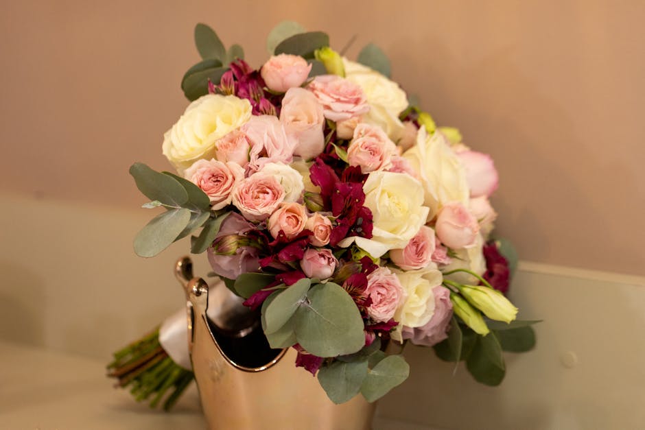 A floral bouquet arranged with pale pink roses, cream roses, and deep burgundy flowers, accented by round, silvery-green eucalyptus leaves. The bouquet displays a variety of textures and sizes, with s
