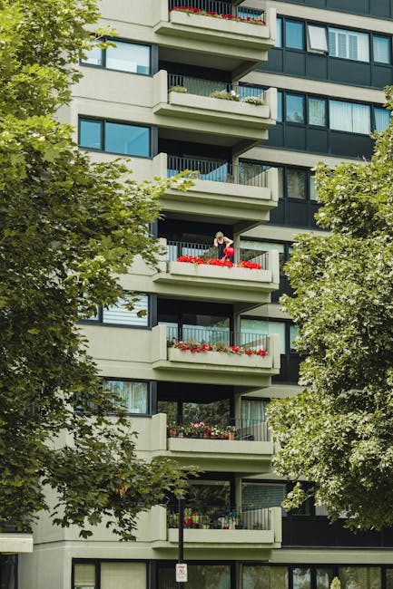 A photograph capturing a multi-storey residential building with several balconies decorated with vibrant flower arrangements. The building's exterior is modern, featuring large windows and light-color