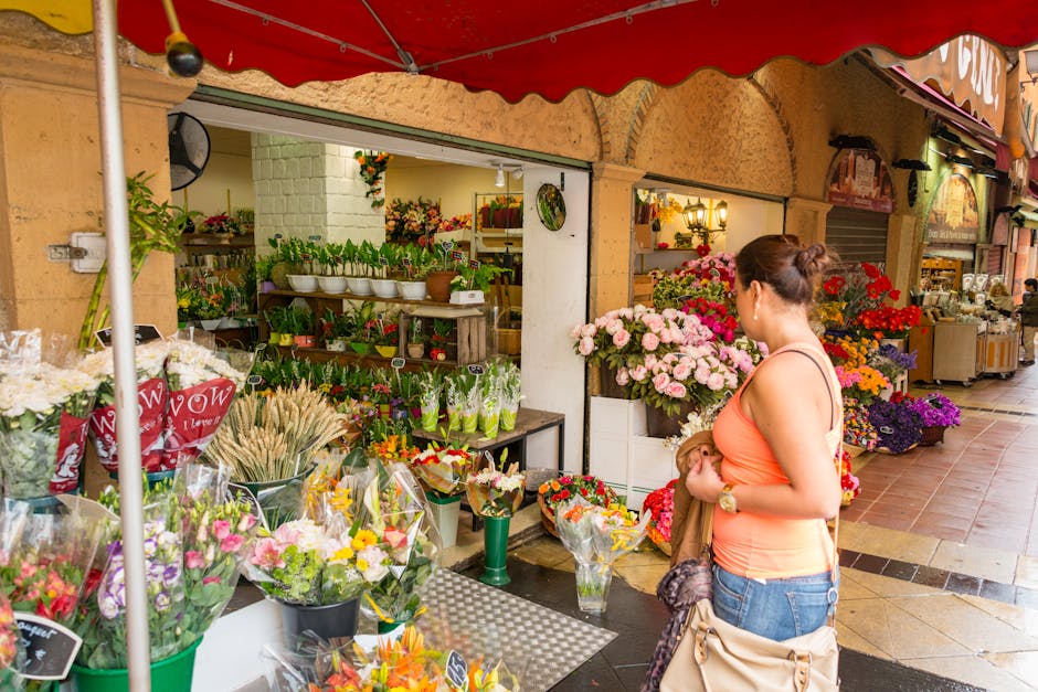 A woman with dark hair tied in a bun, wearing an orange sleeveless top and blue jeans, stands in front of a flower shop under a red canopy at Crystal Palace Park. She is holding a brown coat and a bei